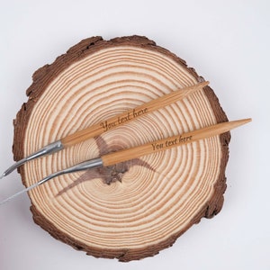 May include: Two wooden knitting needles with the text "You text here" engraved on them. The needles are lying on a wooden surface with a circular grain pattern.