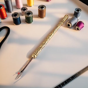 May include: A gold-colored seam ripper with ornate detailing, lying on a white surface. Various spools of thread in colors like pink, blue, and brown are scattered around the ripper. A ruler is visible in the bottom right corner.