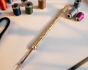 May include: A gold-colored seam ripper with ornate detailing, lying on a white surface. Various spools of thread in colors like pink, blue, and brown are scattered around the ripper. A ruler is visible in the bottom right corner.