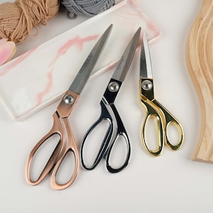 May include: Three pairs of fabric scissors with different colored handles: rose gold, silver, and gold. The scissors are on a white marble surface.
