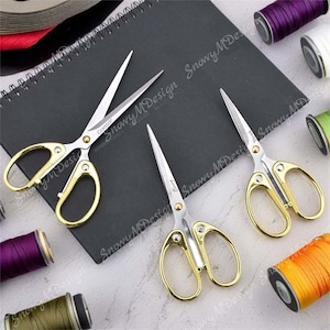 May include: Three pairs of gold-handled scissors with silver blades. The scissors are on a black surface. The text "Caesar" is printed on one of the scissors.