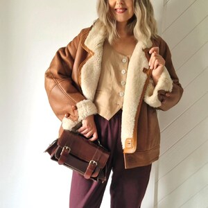 Front view of a model wearing a brown shearling jacket with cream wool lining, visible wide collar and rolled cuffs, button front closure and hip-length cut. The jacket is worn open, showing a light beige buttoned top underneath. The model holds a brown leather handbag and stands indoors against a light wall.