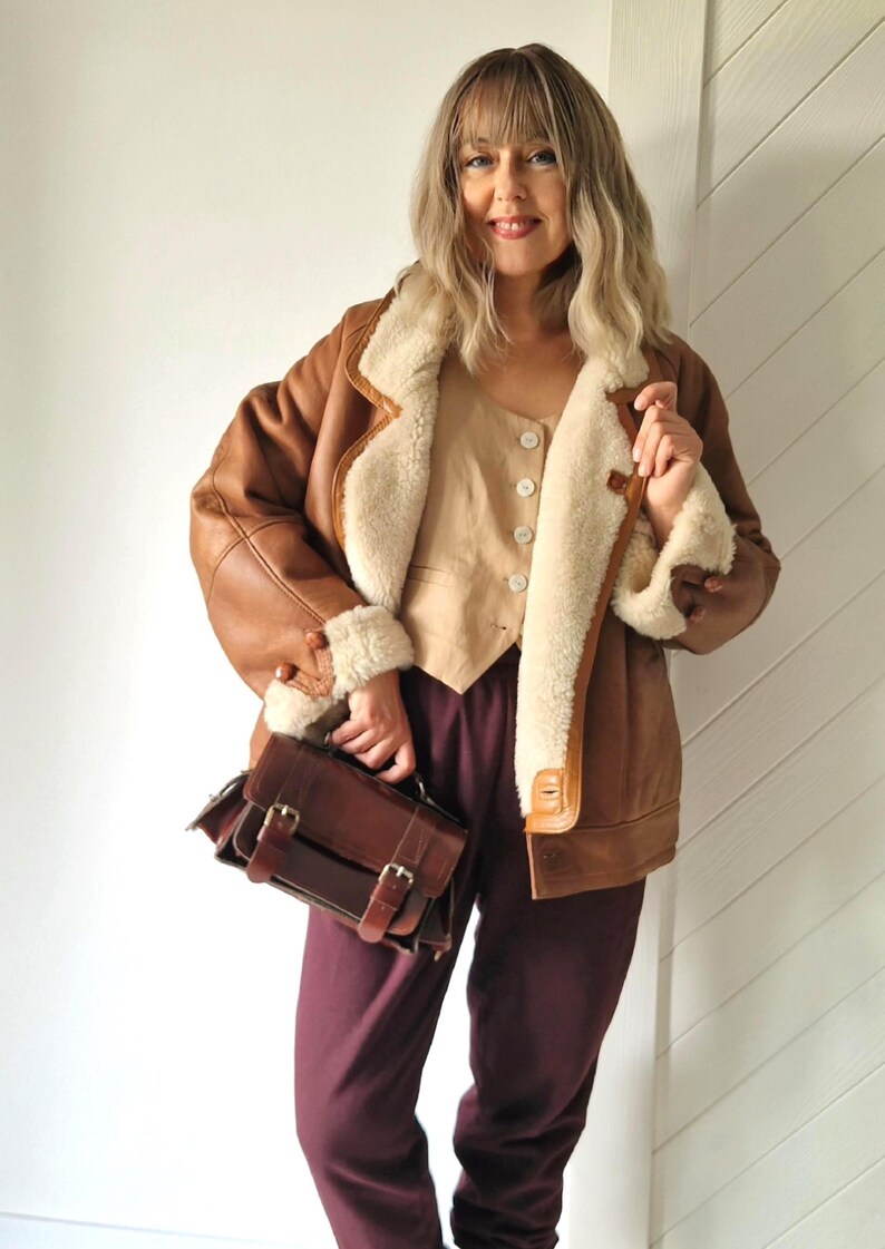 Front view of a model wearing a brown shearling jacket with cream wool lining, visible wide collar and rolled cuffs, button front closure and hip-length cut. The jacket is worn open, showing a light beige buttoned top underneath. The model holds a brown leather handbag and stands indoors against a light wall.