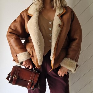 A brown leather jacket with cream-colored real lamb fur, shearling lining and trim. The jacket has a classic design with button closures and a wide collar. A brown leather satchel with buckles is held in the hand.