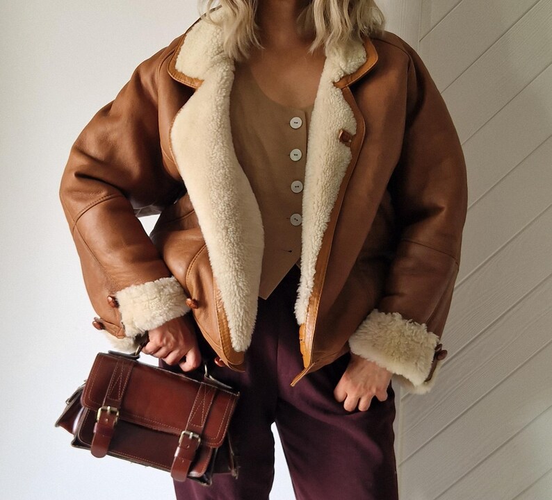 A brown leather jacket with cream-colored real lamb fur, shearling lining and trim. The jacket has a classic design with button closures and a wide collar. A brown leather satchel with buckles is held in the hand.