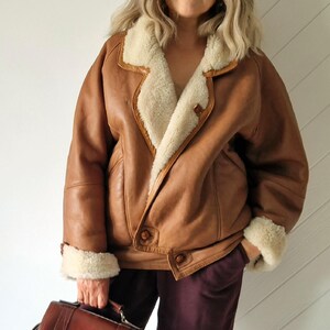 A brown leather jacket with a cream-colored shearling lining and trim. The jacket has a button closure and two front pockets. The cuffs are also trimmed with shearling. A brown leather briefcase is held in the hand.