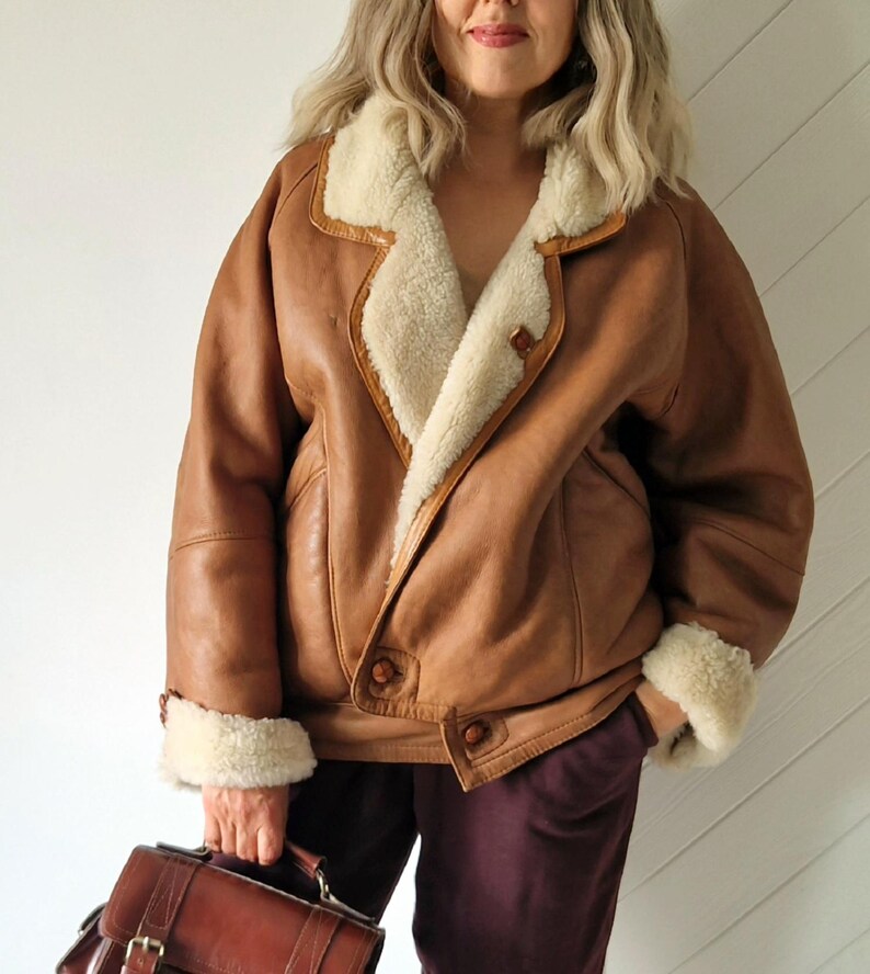 A brown leather jacket with a cream-colored shearling lining and trim. The jacket has a button closure and two front pockets. The cuffs are also trimmed with shearling. A brown leather briefcase is held in the hand.