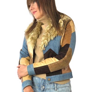 Könnte beinhalten: Eine Patchwork-Jacke mit Einsätzen aus blauem Denim, beigem Wildleder und dunkelbraunem Wildleder. Die Jacke hat einen großen, flauschigen cremefarbenen Kunstfellkragen und lange Ärmel mit geknöpften Manschetten. Sie wird über einem beigen Rollkragenpullover getragen.
