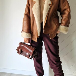 A brown leather jacket with a cream-colored shearling lining and trim. The jacket is paired with burgundy pants, a brown leather satchel, and burgundy pointed-toe heels with buckle details. The outfit is a mix of textures and colors.