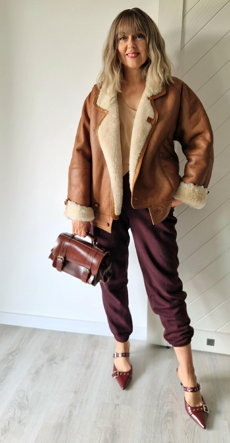 A brown leather jacket with a cream-colored shearling lining and trim. The jacket is paired with burgundy pants, a brown leather satchel, and burgundy pointed-toe heels with buckle details. The outfit is a mix of textures and colors.
