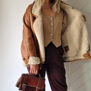 A brown leather shearling jacket with a cream-colored interior and trim. The jacket is paired with a tan vest and burgundy pants. A brown leather satchel bag with buckles is held in the hand.