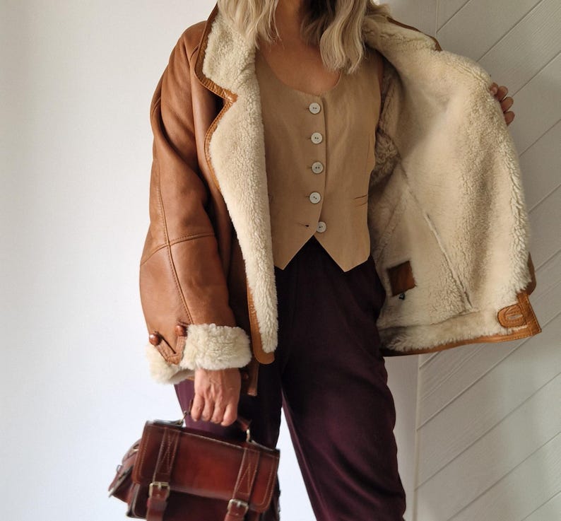 A brown leather shearling jacket with a cream-colored interior and trim. The jacket is paired with a tan vest and burgundy pants. A brown leather satchel bag with buckles is held in the hand.