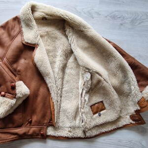 A brown leather jacket with a cream-colored shearling lining. The jacket has a classic design with a notched collar, button closures, and shearling trim on the cuffs. The jacket is open, revealing the soft, warm interior.
