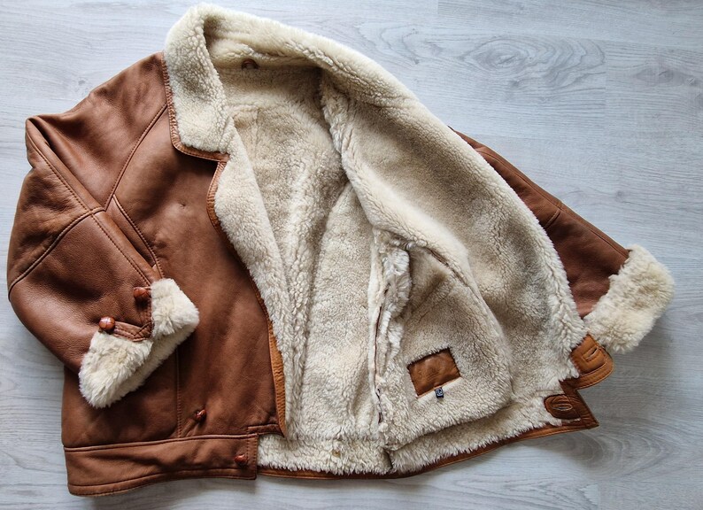 A brown leather jacket with a cream-colored shearling lining. The jacket has a classic design with a notched collar, button closures, and shearling trim on the cuffs. The jacket is open, revealing the soft, warm interior.