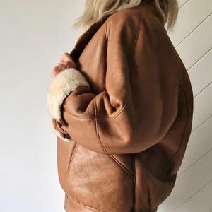 A brown leather jacket with a shearling collar and cuffs. The jacket has a classic bomber style with a ribbed hem and cuffs. The leather appears to be distressed, giving it a vintage look. The jacket is buttoned at the cuffs.