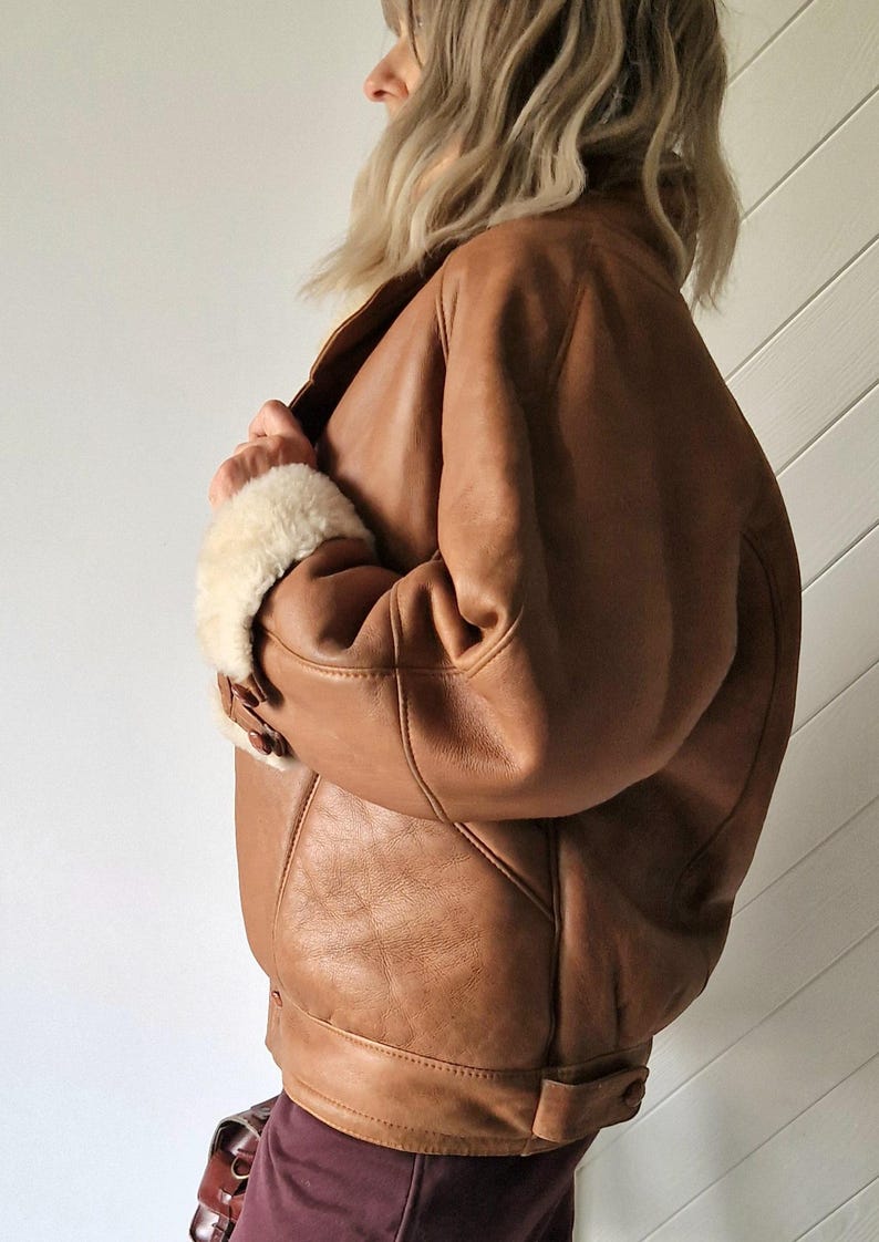 A brown leather jacket with a shearling collar and cuffs. The jacket has a classic bomber style with a ribbed hem and cuffs. The leather appears to be distressed, giving it a vintage look. The jacket is buttoned at the cuffs.