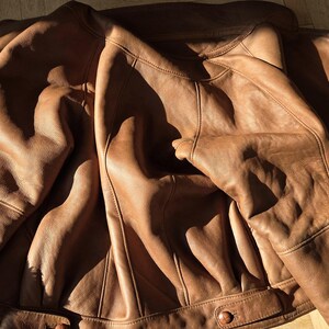 A brown leather jacket with a shearling lining. The jacket is laid out on a wooden floor, with the sunlight casting shadows across the surface. The jacket has a classic design with button closures.