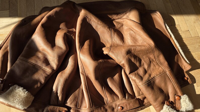 A brown leather jacket with a shearling lining. The jacket is laid out on a wooden floor, with the sunlight casting shadows across the surface. The jacket has a classic design with button closures.