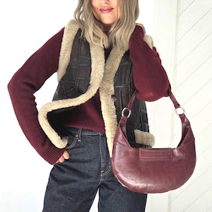 May include: A burgundy leather shoulder bag with silver-tone hardware. The bag has a crescent shape and a flap closure. The person is wearing a burgundy sweater, a dark brown leather vest with cream-colored trim, and dark wash jeans.