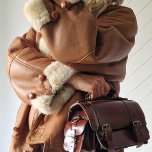 A brown leather jacket with cream-colored shearling trim on the cuffs and collar. The jacket has large buttons and is paired with a matching brown leather satchel bag with buckles. The bag is held by the handle.