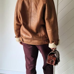 A brown leather jacket with a shearling lining and a matching brown leather handbag. The jacket has a wide collar and cuffs with exposed shearling. The person is wearing burgundy pants and brown heeled sandals.