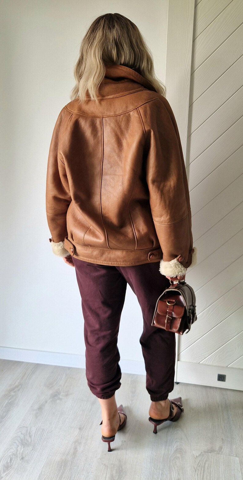 A brown leather jacket with a shearling lining and a matching brown leather handbag. The jacket has a wide collar and cuffs with exposed shearling. The person is wearing burgundy pants and brown heeled sandals.
