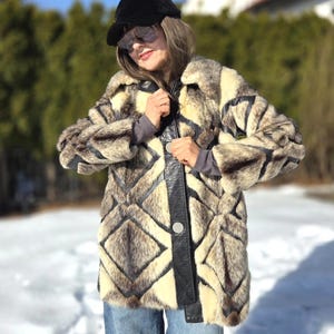 May include: A faux fur coat with a geometric pattern in shades of brown, cream, and black. The coat features a black leather-like trim and a single button closure. The person in the image is wearing a black cap and sunglasses.