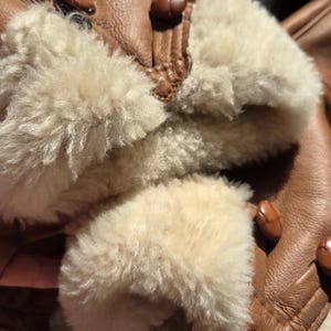 Close-up of a brown leather jacket with cream-colored shearling cuffs. The jacket features brown buttons and a soft, textured interior. The image highlights the jacket's warm, inviting design.