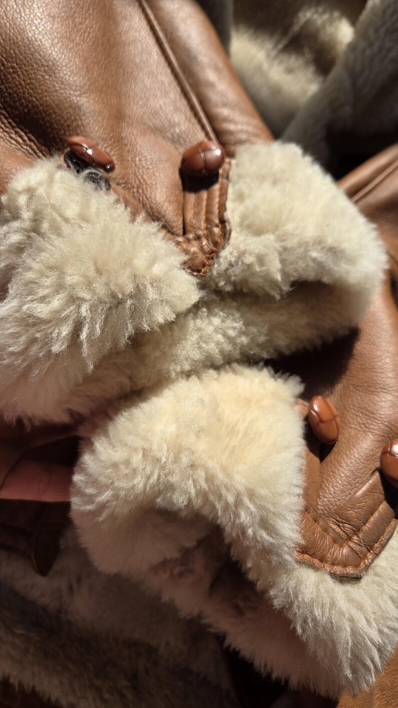 Close-up of a brown leather jacket with cream-colored shearling cuffs. The jacket features brown buttons and a soft, textured interior. The image highlights the jacket's warm, inviting design.