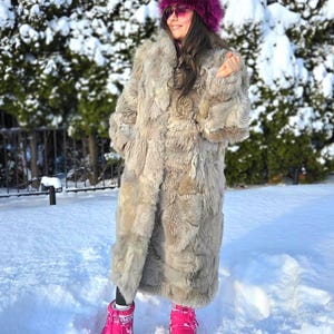 Puede incluir: Un abrigo largo de piel marrón claro se usa con un gorro de piel a juego y botas rosas brillantes. El abrigo llega hasta la rodilla y tiene una textura peluda. El sombrero es de color púrpura oscuro. Las botas son de color rosa vibrante.