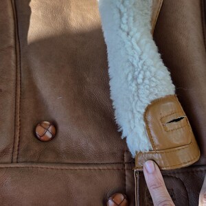 Close-up of a brown leather jacket with a cream-colored shearling collar. The jacket features two brown buttons and a tan leather trim. The texture of the leather is visible.