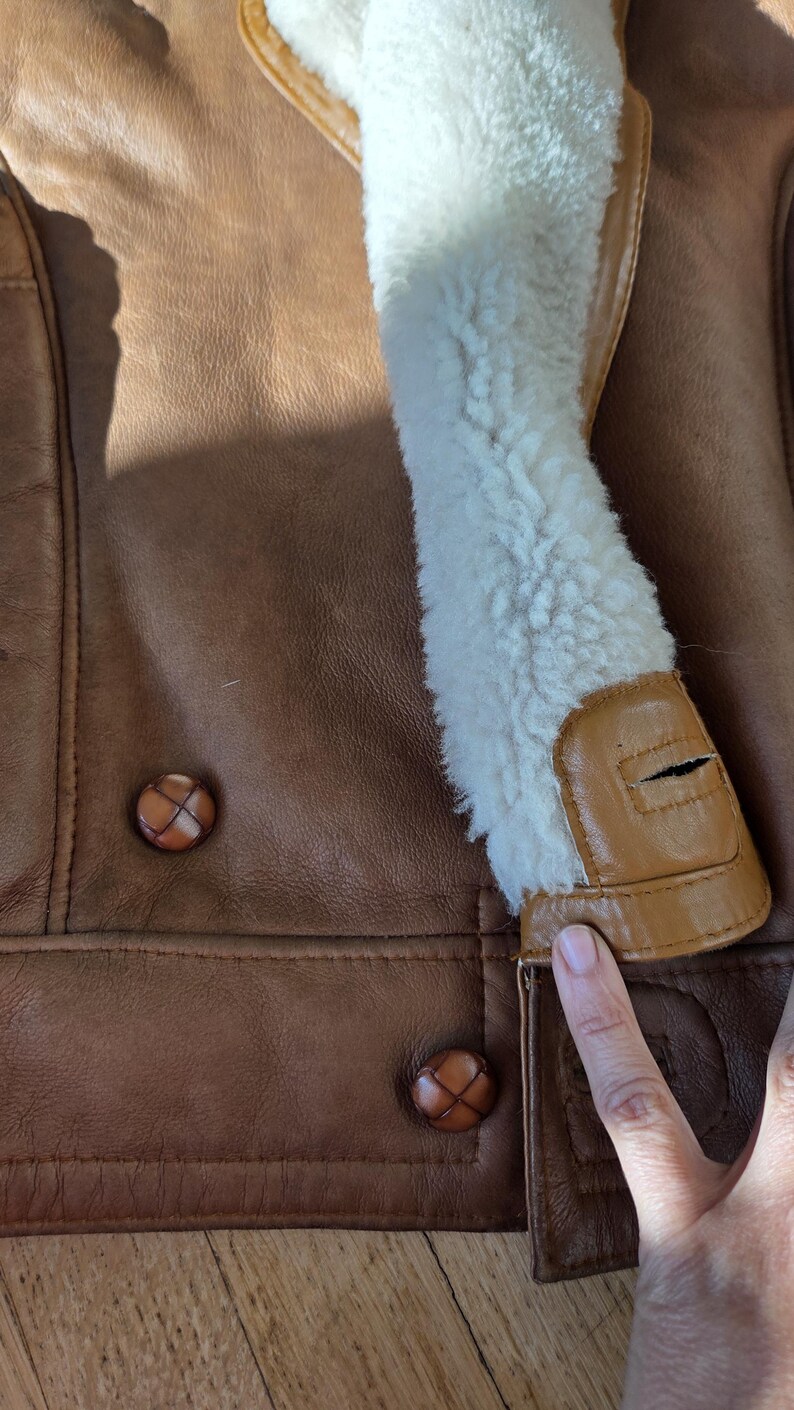 Close-up of a brown leather jacket with a cream-colored shearling collar. The jacket features two brown buttons and a tan leather trim. The texture of the leather is visible.