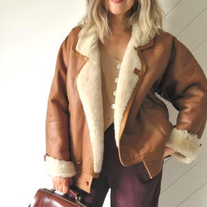 Front view of a model wearing a brown shearling jacket with cream wool lining, visible wide collar and rolled cuffs, button front closure and hip-length cut. The jacket is worn open, showing a light beige buttoned top underneath. The model holds a brown leather handbag and stands indoors against a light wall.