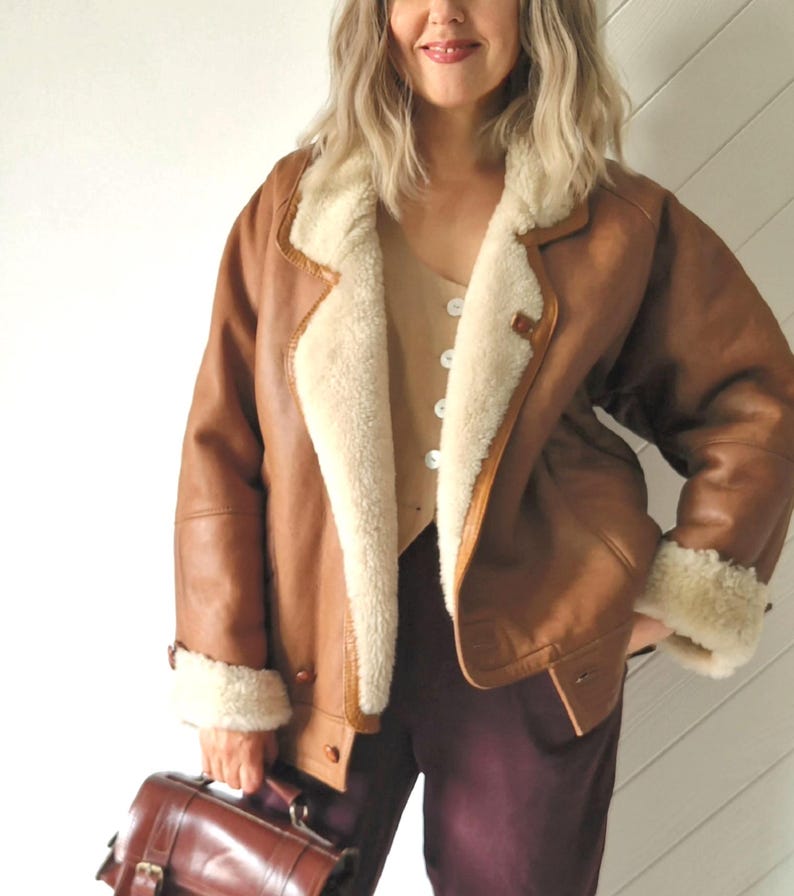Front view of a model wearing a brown shearling jacket with cream wool lining, visible wide collar and rolled cuffs, button front closure and hip-length cut. The jacket is worn open, showing a light beige buttoned top underneath. The model holds a brown leather handbag and stands indoors against a light wall.