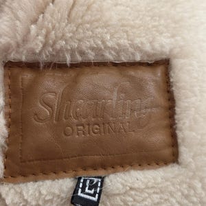 Close-up of a shearling garment with a brown leather label. The label reads "Shearling ORIGINAL" in embossed lettering. The garment has a cream-colored, fluffy interior and a small black tag.