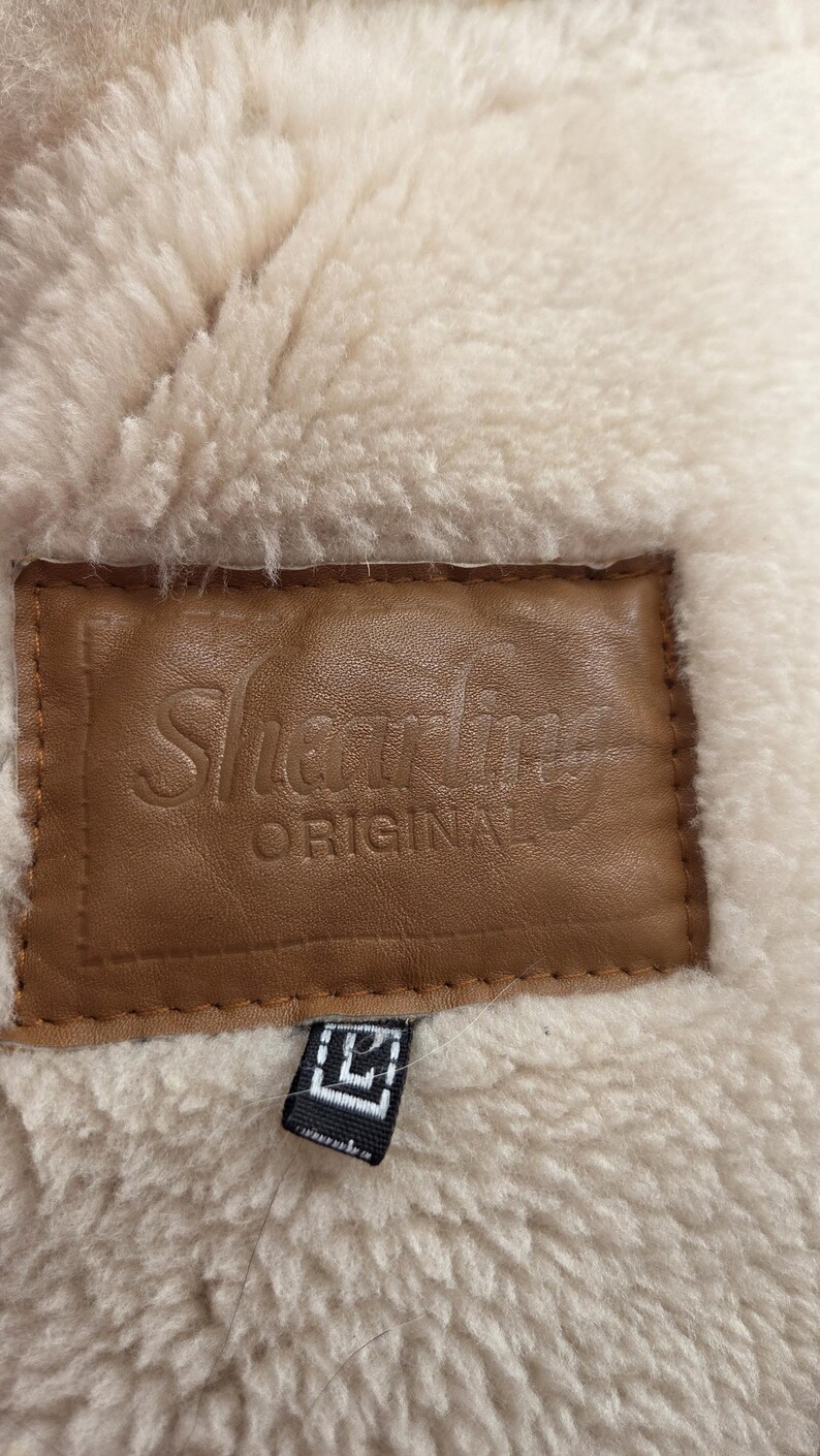 Close-up of a shearling garment with a brown leather label. The label reads "Shearling ORIGINAL" in embossed lettering. The garment has a cream-colored, fluffy interior and a small black tag.