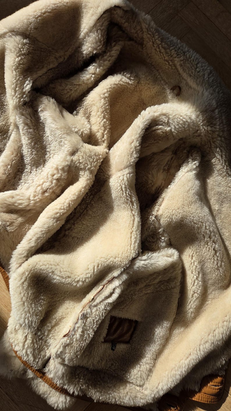 A beige shearling coat with a brown leather trim. The coat has a soft, fluffy interior and a textured exterior. The image shows the coat draped, with details of the stitching and the leather patch.