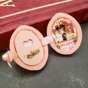 May include: A pink wooden egg-shaped box with a hinged lid. The inside of the box is decorated with a miniature wedding scene with two white bunnies, a cake, and flowers. The lid has a floral design and the words "Happy Wedding" are written on the inside of the box.