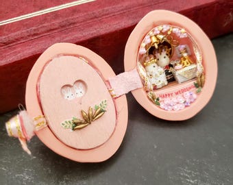 May include: A pink wooden egg-shaped box with a hinged lid. The inside of the box is decorated with a miniature wedding scene with two white bunnies, a cake, and flowers. The lid has a floral design and the words "Happy Wedding" are written on the inside of the box.