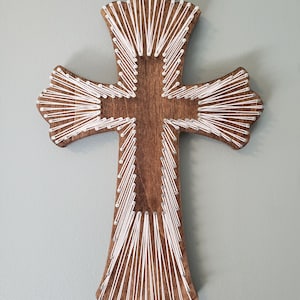 May include: White string art cross on a dark wood background. The cross is made with white string wrapped around nails hammered into the wood. The wood is a dark brown color. The cross is hanging on a light green wall.