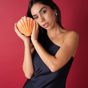 Könnte beinhalten: Eine orangefarbene Clutch in Muschelform mit strukturierter Oberfläche. Die Clutch wird vor einem dunkelblauen Kleid vor rotem Hintergrund gehalten. Die Clutch hat einen perlmuttfarbenen Verschluss.