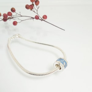 May include: A silver bracelet with a cylindrical charm. The charm has a light blue band with white speckles. A sprig of red berries is in the background.