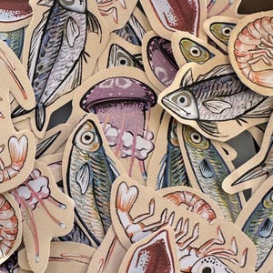 Puede incluir: Una colección de pegatinas troqueladas con ilustraciones de vida marina. Las pegatinas presentan dibujos detallados de peces, camarones, medusas y otras criaturas marinas, con una estética vintage. La paleta de colores incluye tonos de azul, morado y beige.