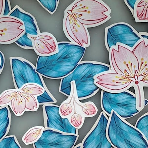May include: An overhead shot of paper cutouts of pink cherry blossoms and teal leaves. The blossoms have pink petals with red outlines and yellow centers. The leaves are teal with dark blue shading. The cutouts are scattered on a gray surface.
