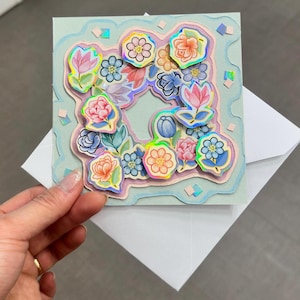 Puede incluir: Una tarjeta de felicitación hecha a mano con un diseño floral. La tarjeta presenta un fondo azul claro con un diseño en capas de pegatinas de flores de colores. La tarjeta se sostiene sobre un sobre blanco.