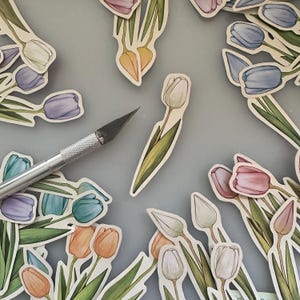 Puede incluir: Una colección de pegatinas troqueladas con ilustraciones de tulipanes en varios colores, incluyendo rosa, morado, naranja, blanco y azul, cada uno con tallos y hojas verdes, dispuestos alrededor de un cúter de metal plateado.