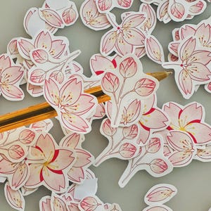 Puede incluir: Una colección de pegatinas de flores de cerezo rosas y blancas con contornos rojos y centros amarillos. Algunas pegatinas son flores abiertas, otras son capullos. Un bolígrafo dorado y negro descansa entre las pegatinas.