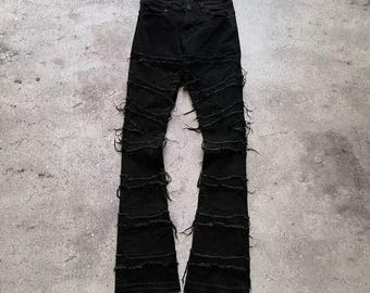 May include: Black denim jeans with a unique distressed design. The jeans feature horizontal frayed detailing across the legs, creating a textured appearance. The pants have a classic five-pocket design and a flared leg opening.
