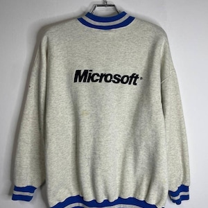 May include: A gray sweatshirt with a blue and white striped collar and cuffs. The sweatshirt has a large black embroidered logo that says "Microsoft".