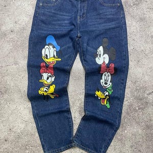 Könnte beinhalten: Blaue Jeans mit Cartoon-Charakter-Grafiken. Die Jeans haben ein klassisches Fünf-Taschen-Design und eine gerade Beinpassform. Die Grafiken zeigen Donald Duck, Daisy Duck, Mickey Mouse, Minnie Mouse und Pluto.